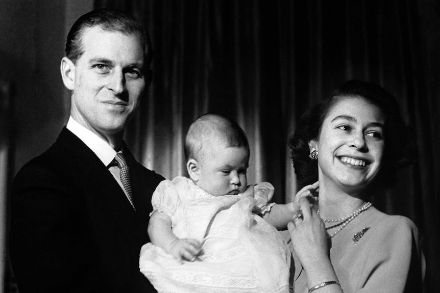Prince Phillip cradles a baby Prince Charles, with Queen Elizabeth II playing with his hand.
