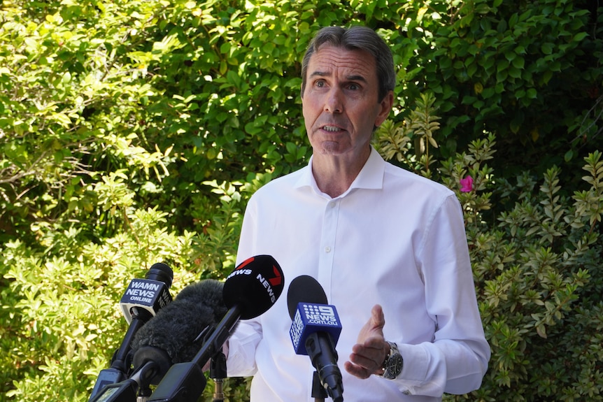 Mid-shot of Peter Collier wearing a white business shirt, speaking in front of media microphones