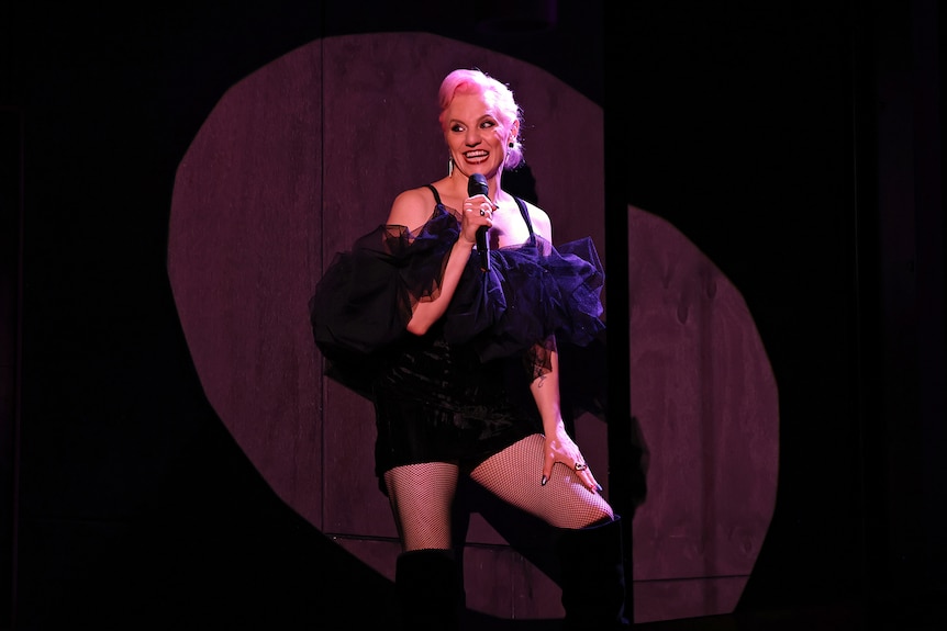 A 30-something trans woman with pink hair, wearing a black dress, talks into a microphone while illuminated by a spotlight