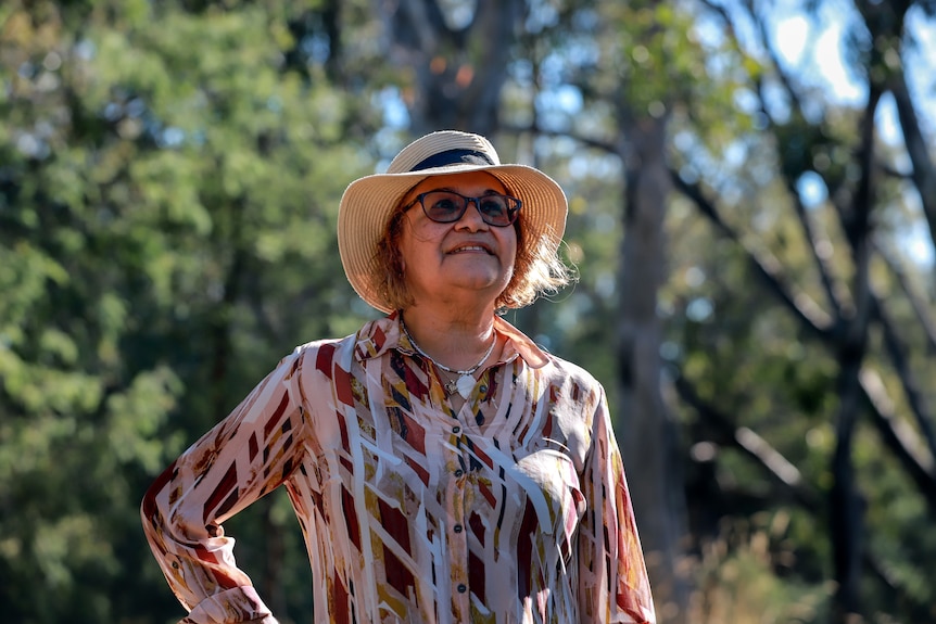 Woman with dark skin wearing hat stands outside with trees behind her.