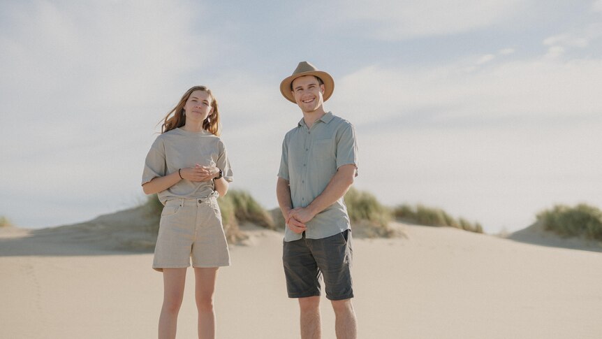 A young woman standing next to a young man wearing a hat, on a sand dune.
