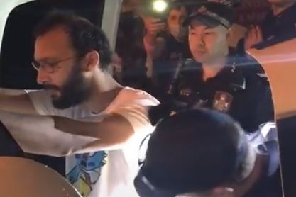Jonathan Sri leans on a car as a police officer appears to check his pockets. Officers and protesters are there.