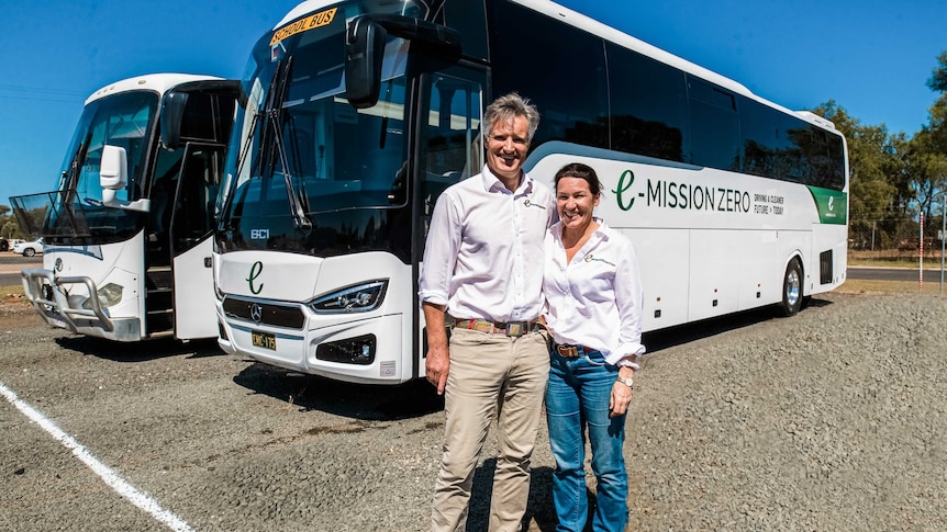 A man and a woman stand together. There is two buses in the background