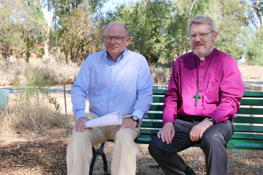 Two men seated on a park bench, one wearing a blue shirt, one wearing a purple shirt with a  priests collar, wearing a cross.