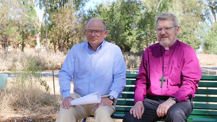 Two men seated on a park bench, one wearing a blue shirt, one wearing a purple shirt with a  priests collar, wearing a cross.