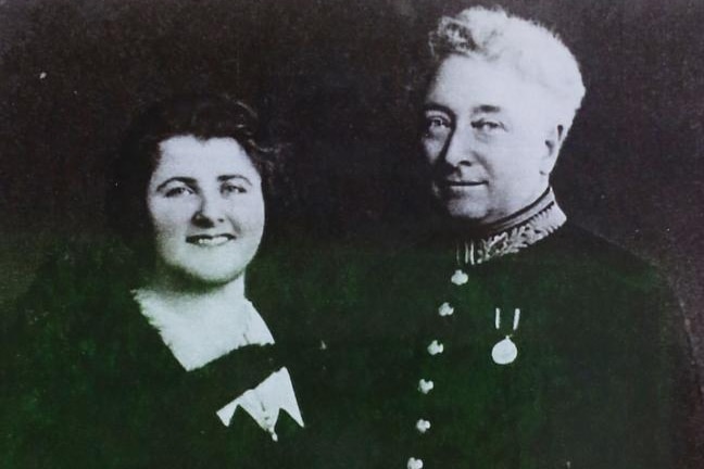 A black and white photo of a white-haired man in a military coat beside a woman smiling broadly. 