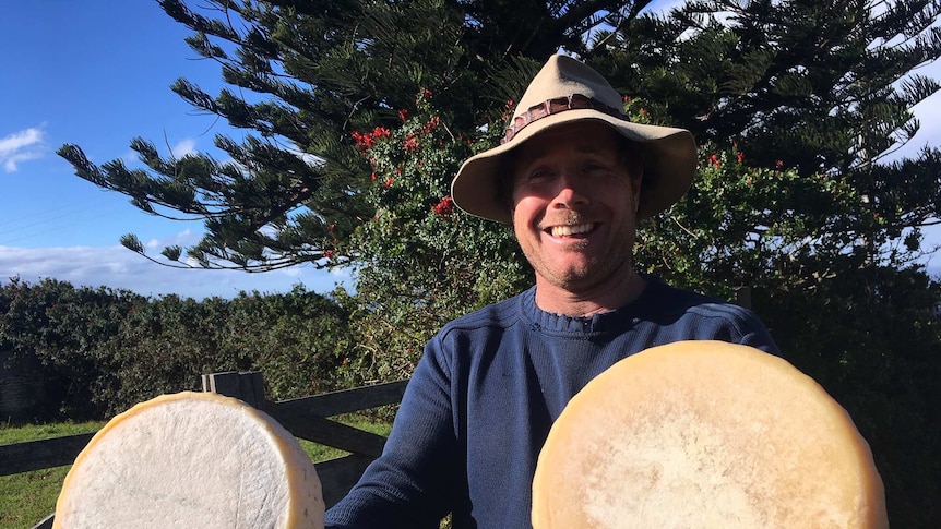 A man in an Akubra-style hat stands smiling holding two wheels of cheese.