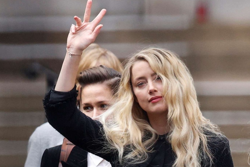 A woman with blond hair, wearing a black jacket waves to a crowd.