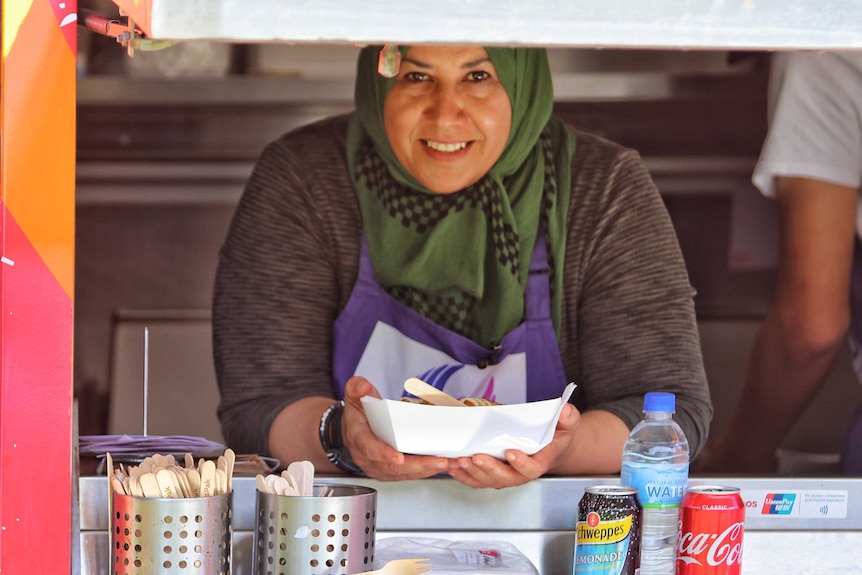 Iraqi refugee Fadhaa Al-Khalidi leans out of the window of her Fare Go food truck holding a tray and smiling.