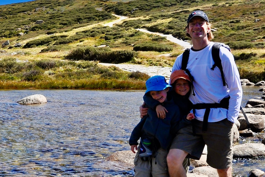 Jono Lineen, with his two young sons, stands in the middle of a creek with bush surrounding them, smiling.