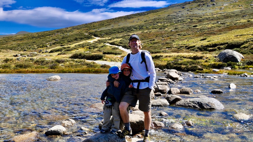 Jono Lineen, with his two young sons, stands in the middle of a creek with bush surrounding them, smiling.