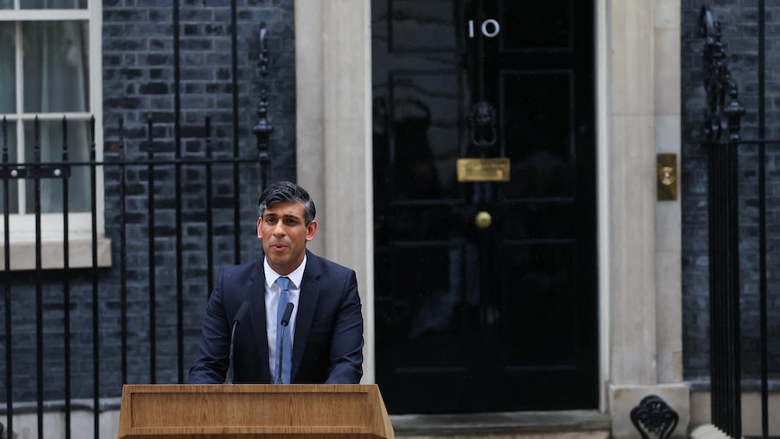 A man in a blue suit stands speaking behind a lectern in the street in front of a black door