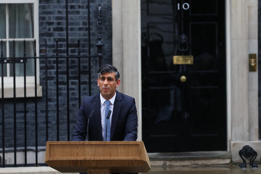 A man in a blue suit stands speaking behind a lectern in the street in front of a black door