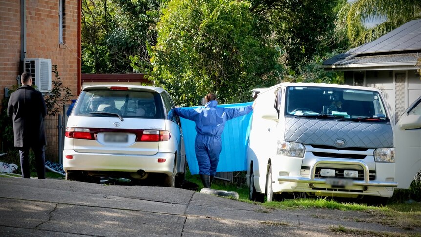 A person in a hazmat suit stands between two cars in a driveway, with a burnt out house in the background.