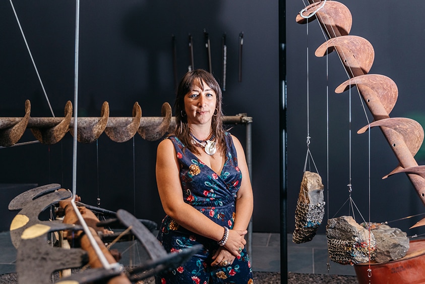 A woman with long dark hair, shell necklace and blue playsuit stands among sculptures made large rust coloured drill augers.