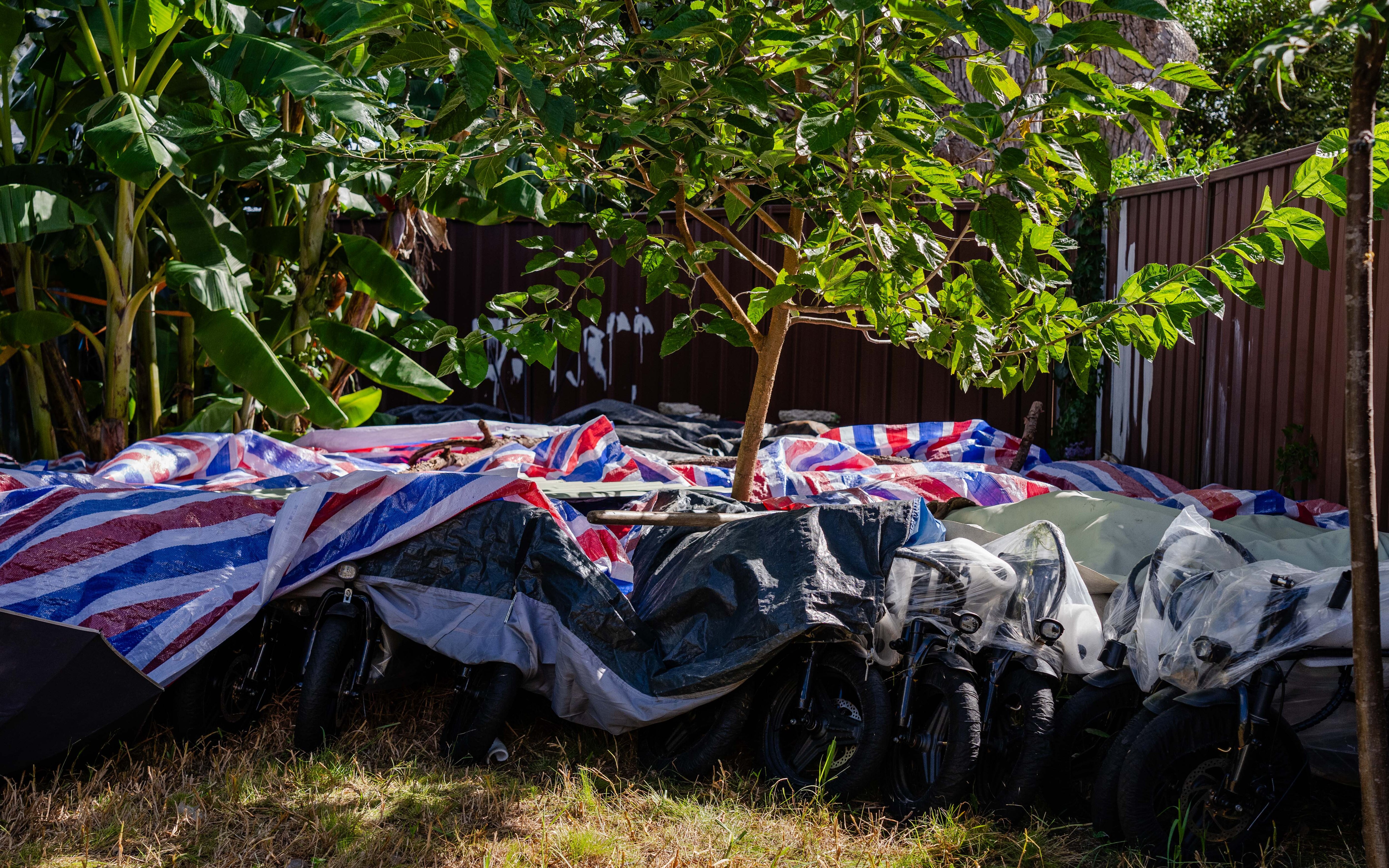 A man in a black shirt and glasses stands in a backyard with e-bikes, some under tarp and some not.
