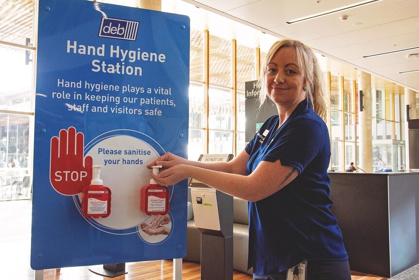 A nurse in a dark blue uniform at a hand sanitiser station inside a hospital foyer