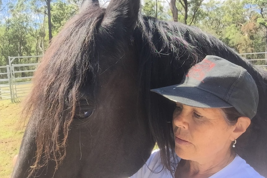 A woman wearing a cap and a white t-shirt stands close to a  horse.