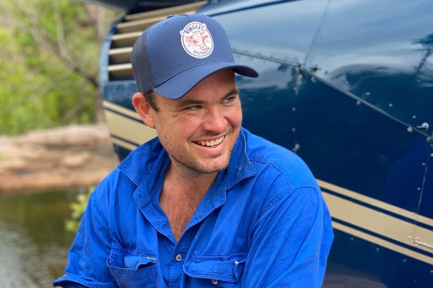Un hombre sonriente sentado en el peldaño de un helicóptero en un prado, con una gorra de camionero y una camisa azul brillante de trabajador agrícola.