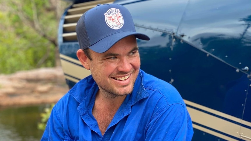 A smiling man sitting on the rung of a helicopter in grassland, wearing a trucker cap and bright blue ag worker shirt.