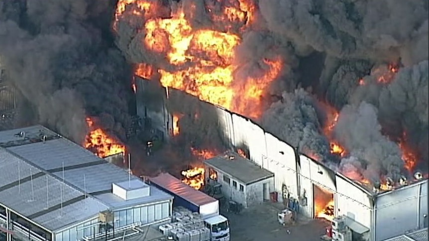 Flames and thick black smoke engulf a large warehouse building.
