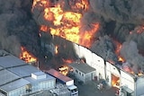 Flames and thick black smoke engulf a large warehouse building.