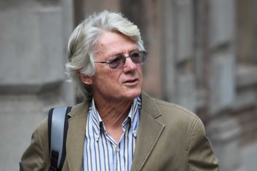 Mid-shot of a grey haired man in glasses walking along a city street, background blurred.