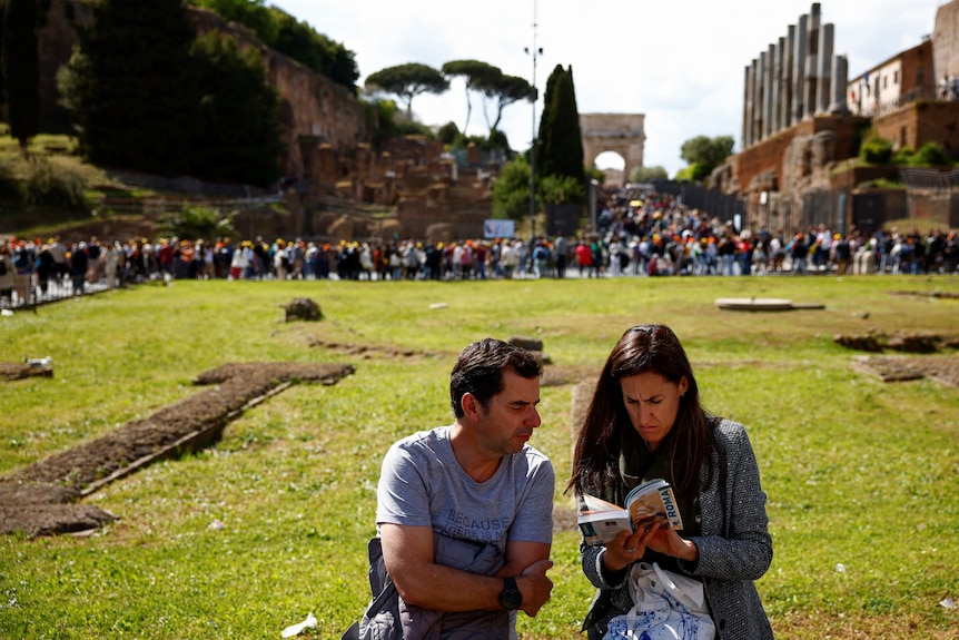 Tourists read a travel guide of Rome as they sit near Colosseum crowds in Rome.