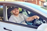 A smiling middle-aged man, wears cap, red beard and moustache, sits in the drivers seat of a car.