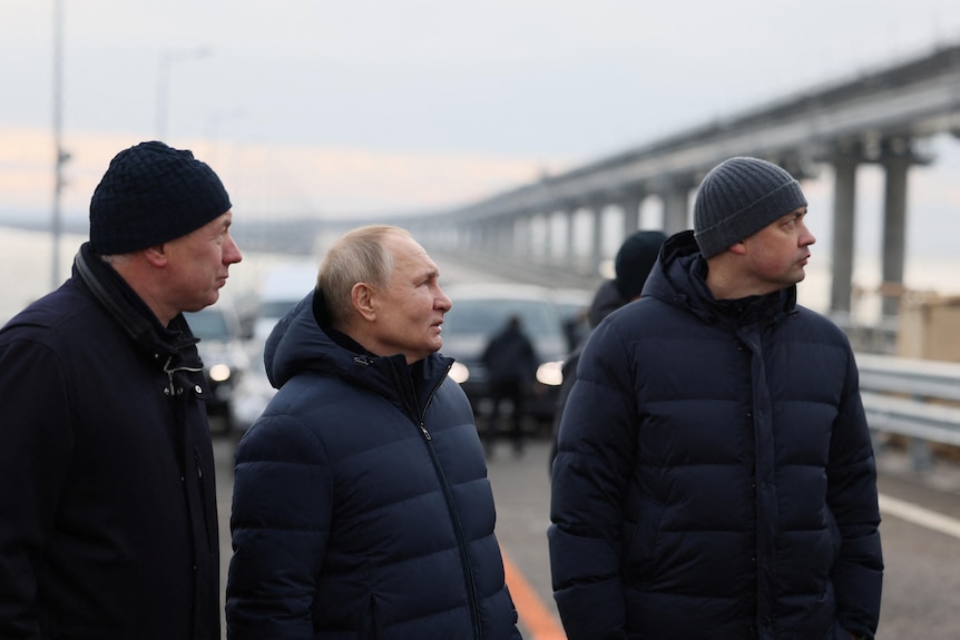 Vladimir Putin stands in a puffer coat with two other men on a road bridge, glancing towards the distance