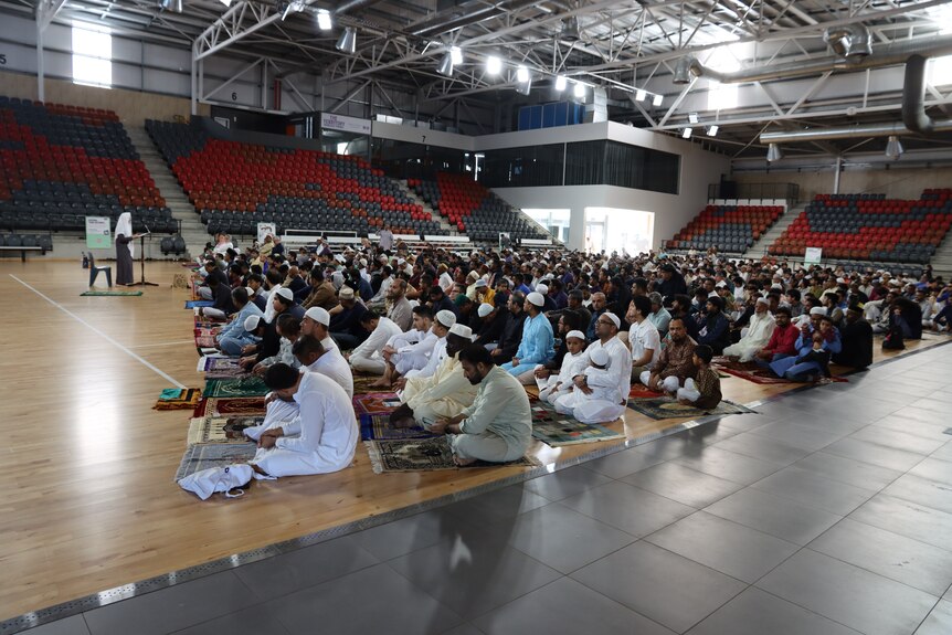 A group of people sitting on mats, some of them praying, inside an indoor sports stadium.
