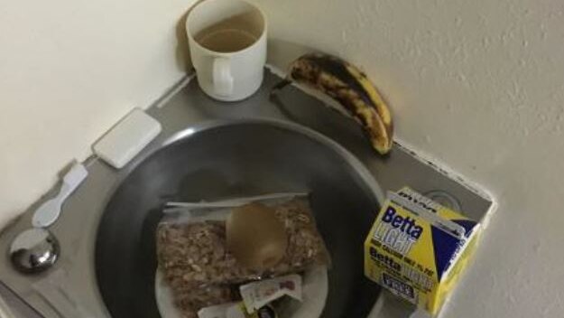A prison cell toilet being used as a table for a cup, banana, milk carton and other breakfast items.