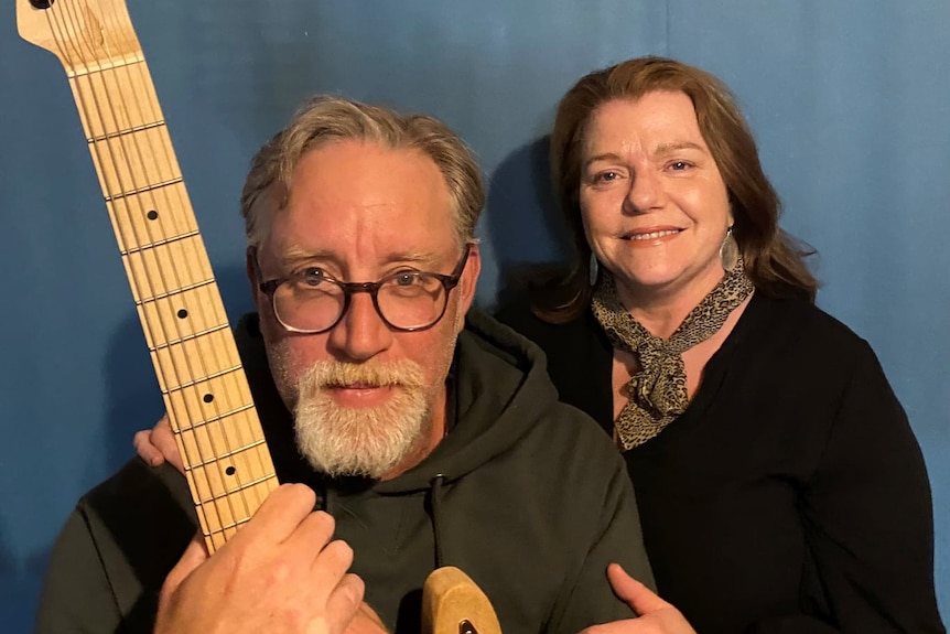 A man with glasses holding a guitar and a woman smiling at the camera. 