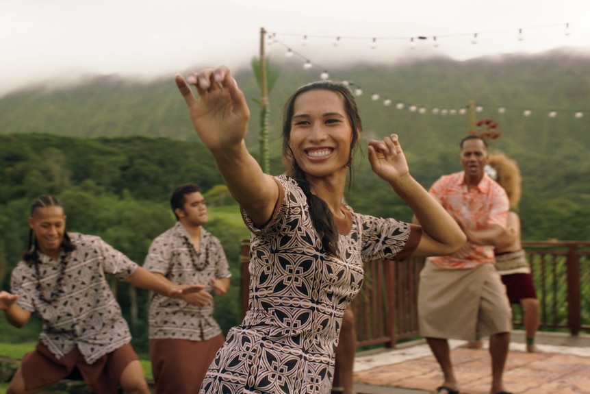 A film still of Kaimana, a fa'afafine actor, dancing and smiling broadly, with other actors from the ensemble dancing behind her