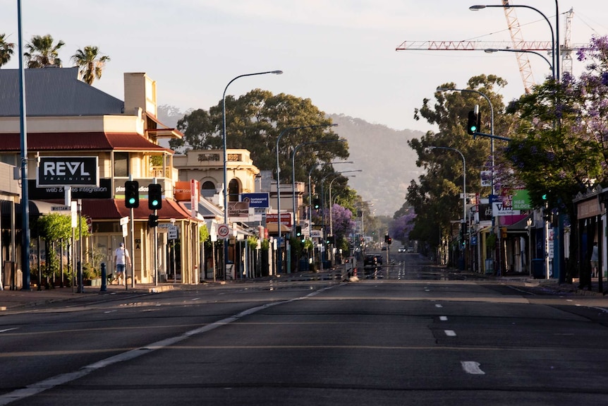 A shop-lined street with hills in the background is nearly empty of cars.