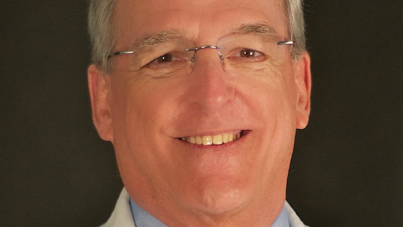 A head and shoulders profile shot of a smiling Quentin Van Meter wearing a white medical coat and a shirt and tie with glasses.