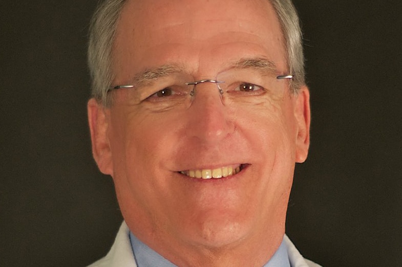 A head and shoulders profile shot of a smiling Quentin Van Meter wearing a white medical coat and a shirt and tie with glasses.
