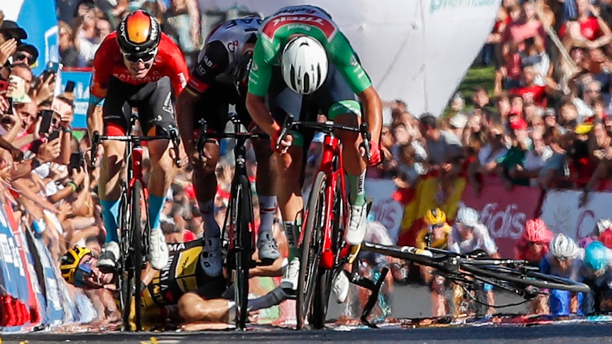 A cyclist in a green jersey puts his head down and sprints ahead at Vuelta, as behind him another cyclist lies on the ground.