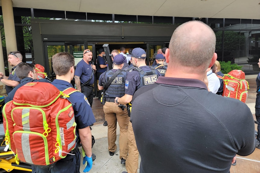 Armed police, one holding a large metal pole, stand near the entrance of a medical building. Medics with large backpacks nearby.