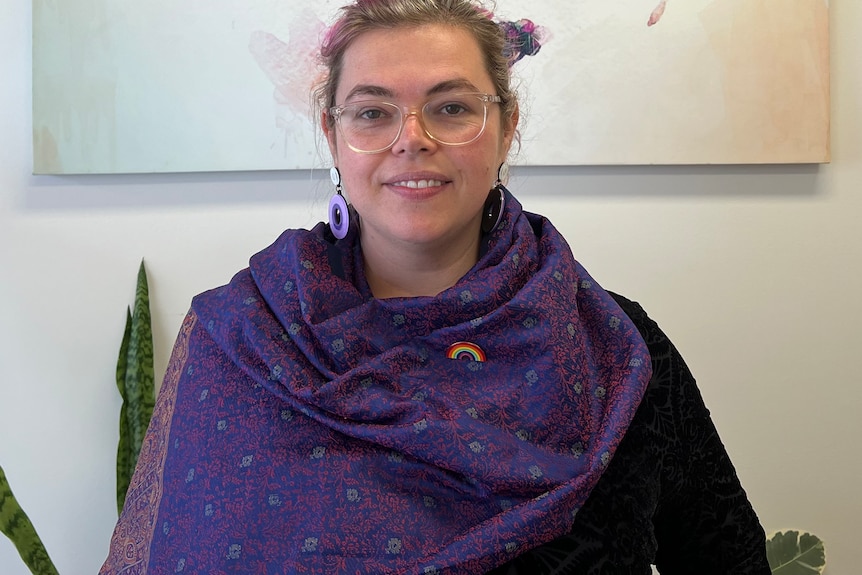 A bespectacled woman with her hair in a bun sits in an office, wearing a shawl.