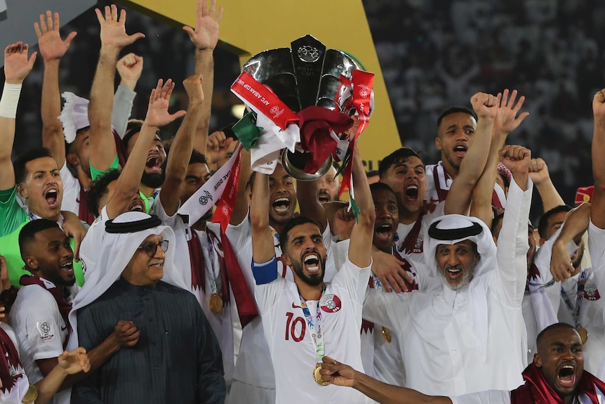 Qatar's players, wearing white kit and standing on a stage, lift a trophy into the air