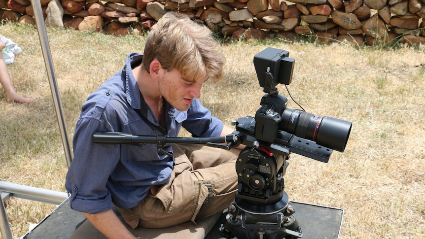 A man in a blue shirt sits on a platform and stares into the back of a mounted camera