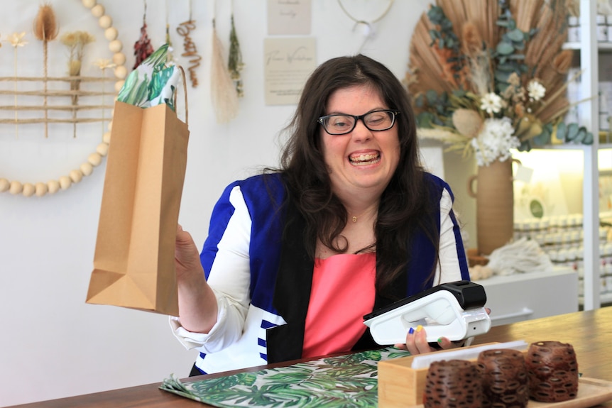 Simone wears a hot pink shirt, blue & white blazer & black glasses, standing behind a shop counter holding a brown, paper bag an