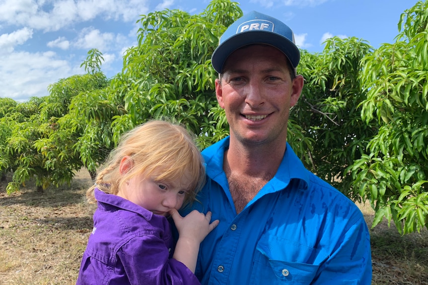 A smiling man holding a little girl who is biting her thumb, with mango trees behind them