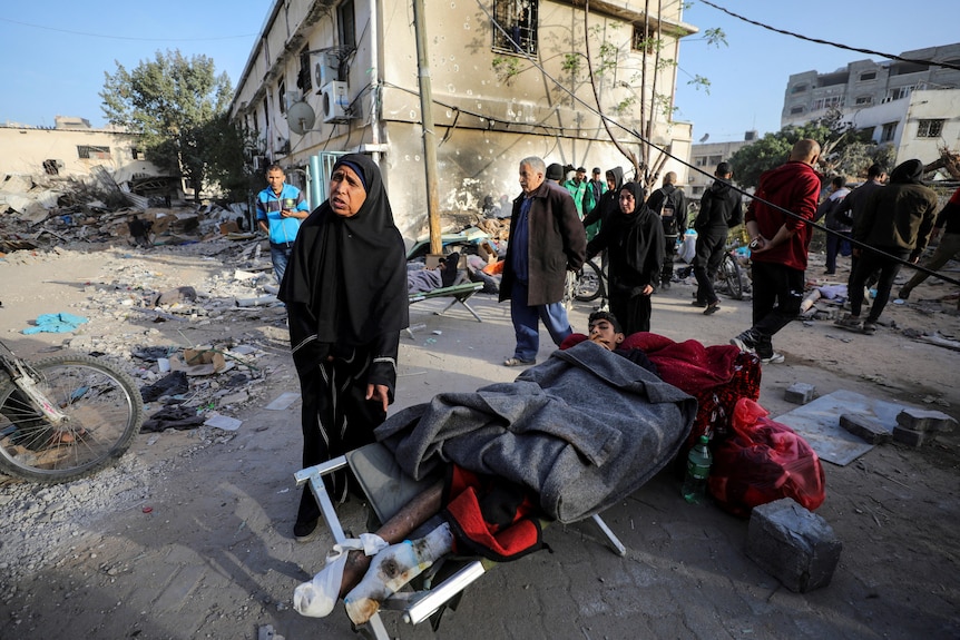 A woman standing next to an injured boy laying on a stretcher outside with rubble on the ground