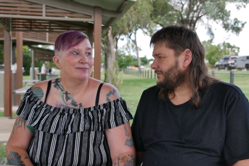 A man and woman sit next to each other at a picnic table in a park and look into each other's eyes.