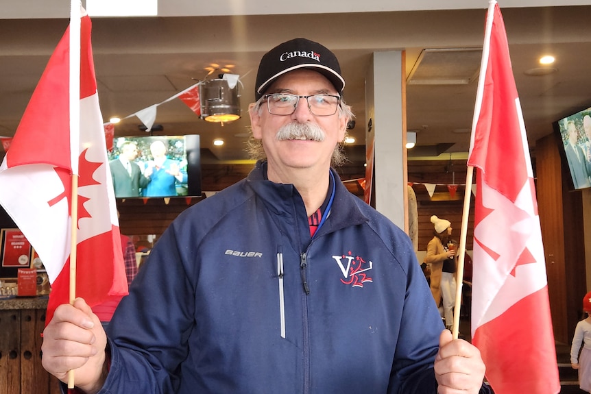 A man wearing glasses, sporting a mustache, holds two Canadian flags and smiles at the camera.