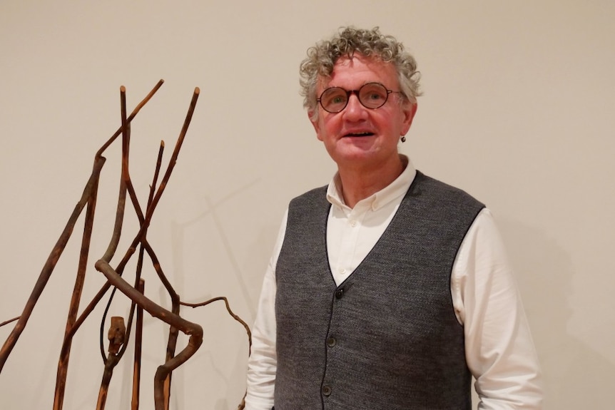 A man in a grey vest next to an wooden art installation looks direct at the camera against a white wall. 