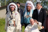 Pope in wheelchair pushed beside men in traditional Indigenous headress.