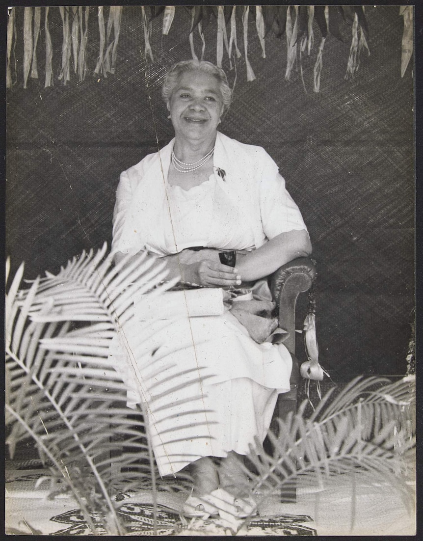 Queen Sālote sits on a chair smiling, presumably to an audience. Decorations of coconut palm branches surround.